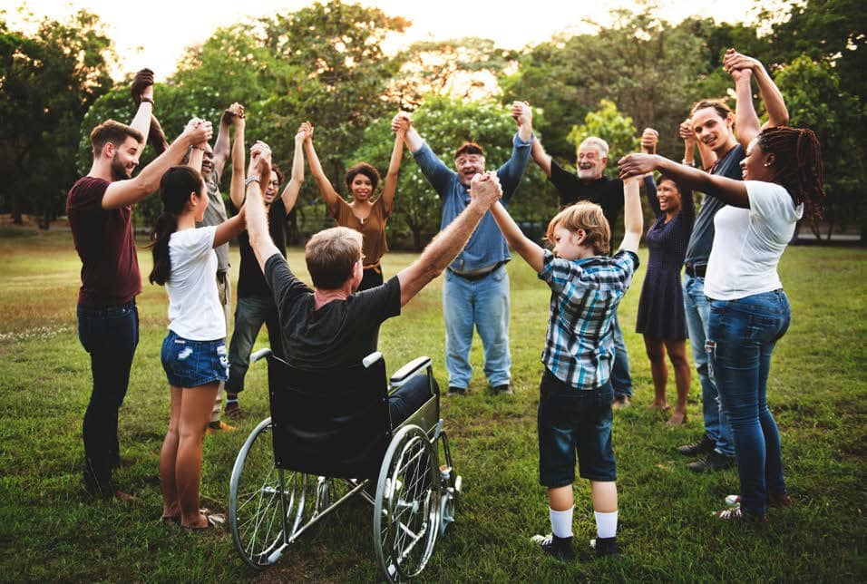 People raising hands in circle together in a field of grass, everyone standing except one man in a wheelchair